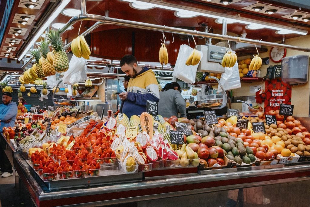 A food stall in La Boqueria Market selling tropical fruits. A bearded man wearing a yellow, white and blue hoodie looking at his phone stands behind the fruits on display.