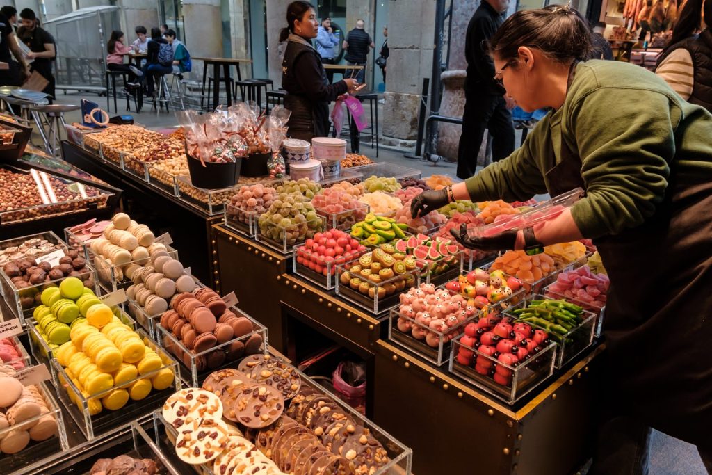 A candy seller arranging candies neatly in La Boqueria Market. Bins of neatly stacked macarons, chocolates, gummies, and all sorts of colorful candy on display.