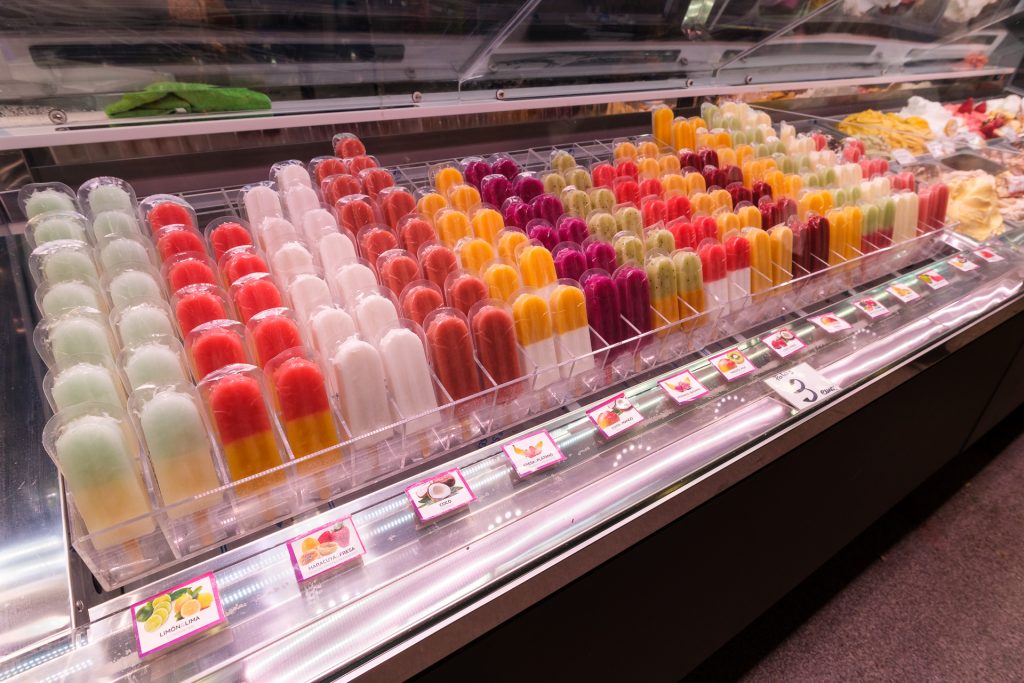Fruit-flavored popsicles of different colors on display in a refrigerated case for sale at La Boqueria Market.